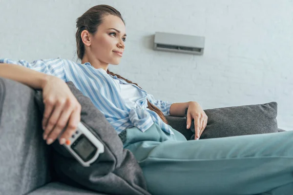 Femme reposant sur canapé avec climatiseur télécommande, chaleur d'été — Photo de stock