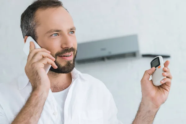 Hombre barbudo hablando en el teléfono inteligente mientras enciende el aire acondicionado con control remoto - foto de stock