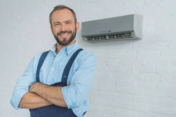 Reparador barbudo sonriente posando con brazos cruzados, con aire acondicionado en la pared - foto de stock