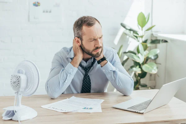 Homem de negócios cansado sentado no escritório com ventilador elétrico, laptop e documentos — Fotografia de Stock