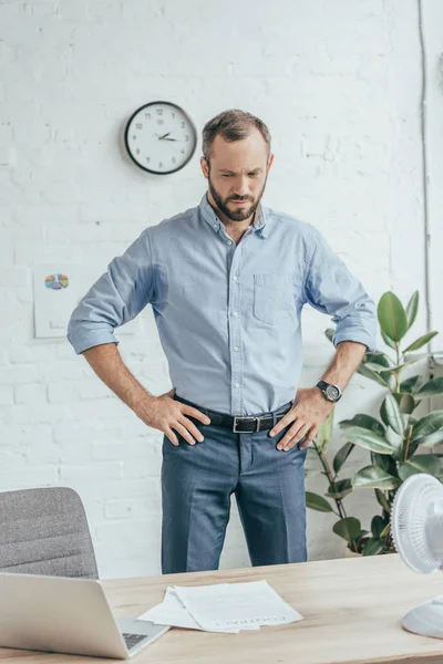 Sweaty businessman in wet shirt standing in office with laptop, documents and electric fan — Stock Photo