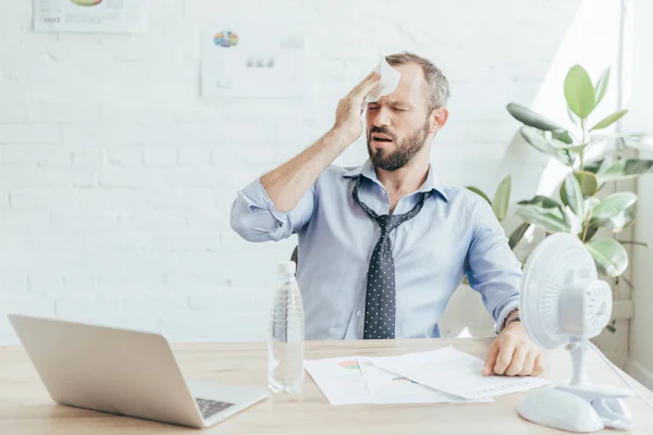 Hombre de negocios cansado con dolor de cabeza sentado en el lugar de trabajo con ventilador eléctrico, botella de agua, papeleo y portátil - foto de stock