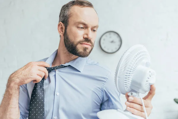 Hombre de negocios quitando corbata y soplando sobre sí mismo con ventilador eléctrico blanco - foto de stock