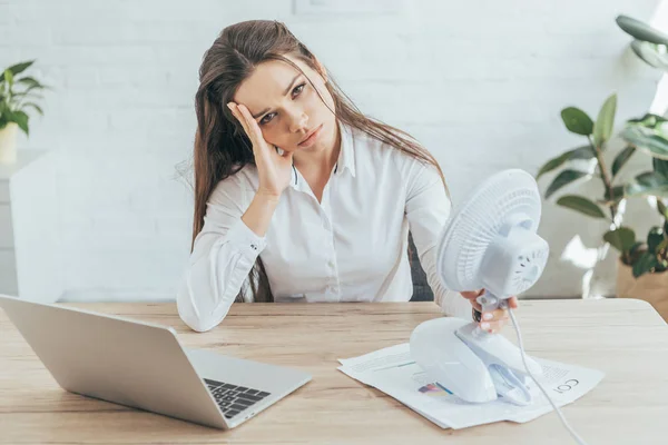 Mulher de negócios chateado sentado no local de trabalho com papelada, laptop e ventilador elétrico — Fotografia de Stock