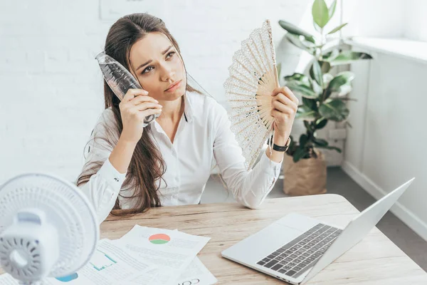 Businesswoman cooling herself with electric fan, hand fan and bottle of water at workplace with documents and laptop — Stock Photo