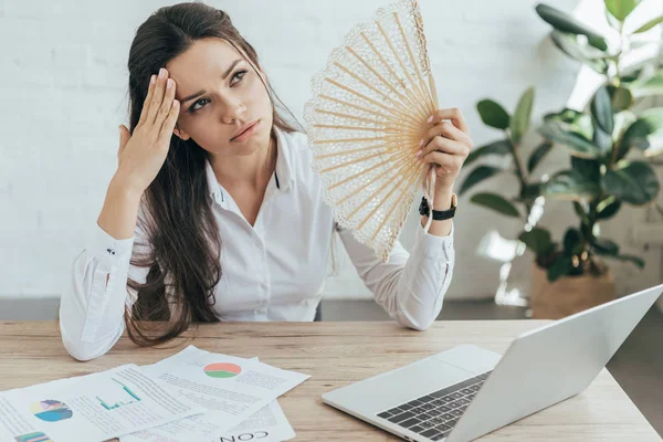 Businesswoman in hot office with laptop and documents blowing herself with hand fan — Stock Photo