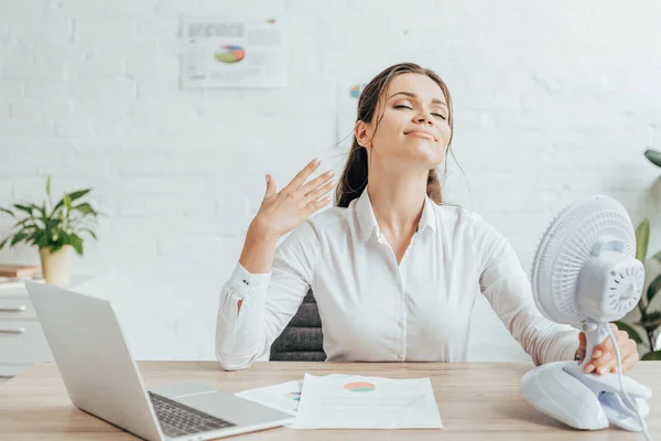 Businesswoman blowing at herself with electric fan while sitting at workplace with documents and laptop — Stock Photo