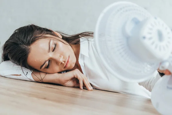 Mujer de negocios agotada con los ojos cerrados soplando en sí misma con ventilador eléctrico en la oficina - foto de stock