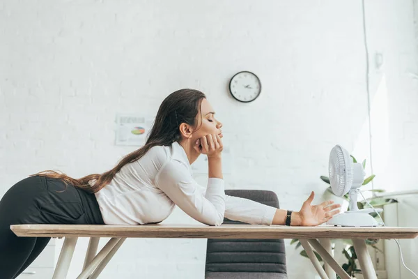 Mujer de negocios soplando en sí misma con ventilador eléctrico en la oficina caliente - foto de stock