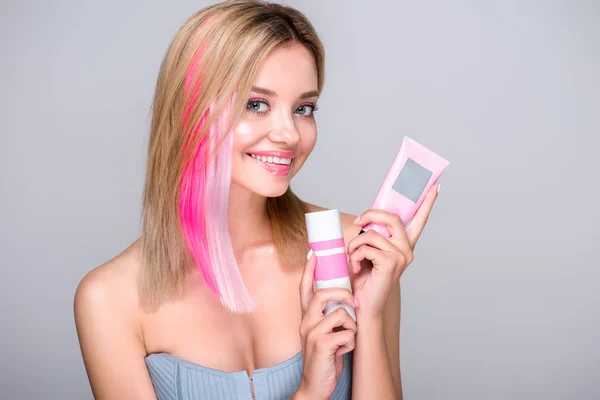 Smiling young woman with colored bob cut holding hair care supplies and looking at camera isolated on grey — Stock Photo