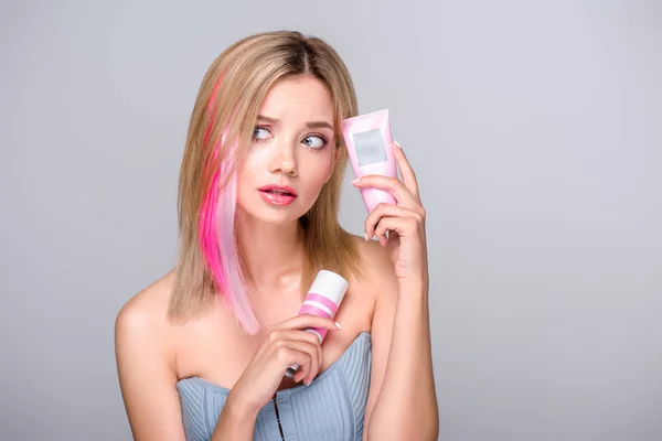 Mujer joven desconcertada con corte bob de color sosteniendo suministros de cuidado del cabello aislados en gris - foto de stock