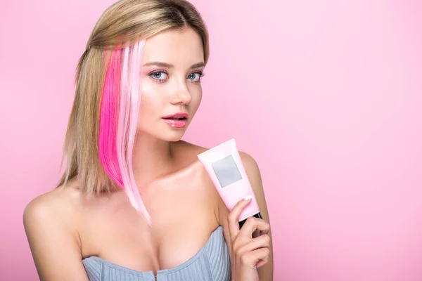 Hermosa mujer joven con hebras de pelo de colores que sostienen el tubo de máscara de pelo y mirando a la cámara aislada en rosa - foto de stock