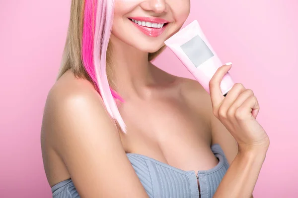 Tiro recortado de mulher jovem sorridente com fios de cabelo coloridos segurando tubo de máscara de cabelo isolado em rosa — Fotografia de Stock