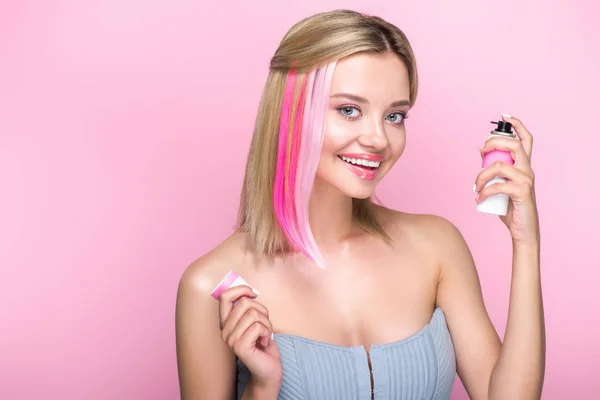 Sonriente mujer joven con hebras de colores de pelo y pintura en aerosol mirando a la cámara aislada en rosa - foto de stock