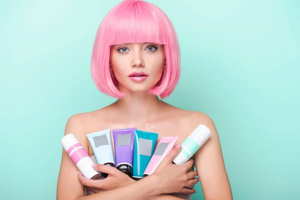 Young woman with pink bob cut holding various tubes of coloring hair tonics looking at camera isolated on turquoise — Stock Photo