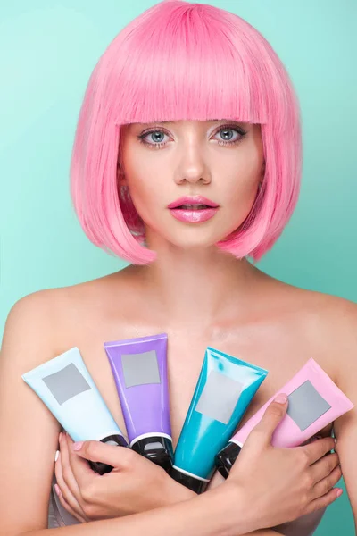 Attrayant jeune femme avec bob rose coupé tenant divers tubes de coloration toniques de cheveux en regardant la caméra isolée sur turquoise — Photo de stock