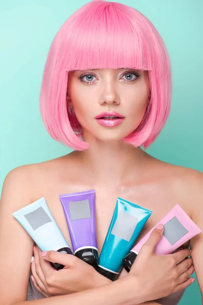 Atractiva mujer joven con corte de color rosa bob celebración de varios tubos de tónicos para colorear el cabello mirando a la cámara aislada en turquesa - foto de stock