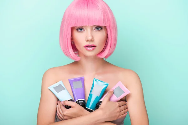 Beautiful young woman with pink bob cut holding various tubes of coloring hair tonics looking at camera isolated on turquoise — Stock Photo