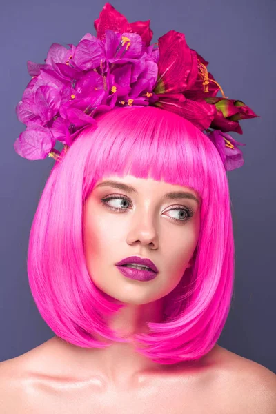 Atractiva mujer joven con corte de color rosa bob y flores en el pelo mirando hacia otro lado aislado en violeta - foto de stock