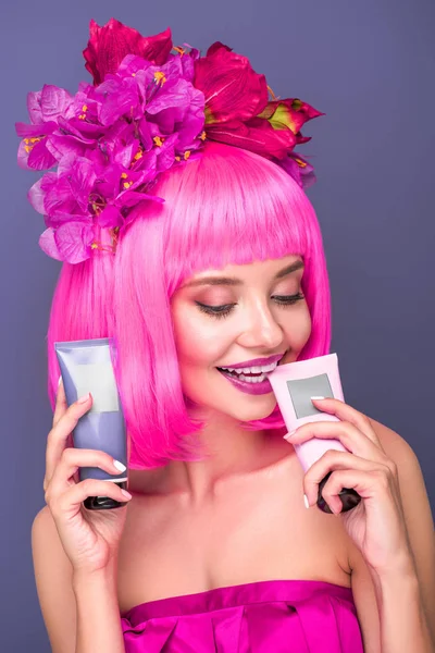 Sonriente mujer joven con el bob rosa cortado y flores en el pelo sosteniendo tubos de tónicos para colorear el cabello aislado en violeta - foto de stock