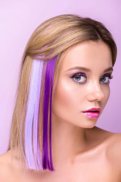 Retrato de cerca de mujer joven atractiva con maquillaje elegante y hebras de pelo púrpura mirando a la cámara aislada en púrpura - foto de stock