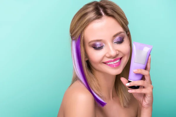 Smiling young woman with colorful hair holding hair tonic tube isolated on blue — Stock Photo