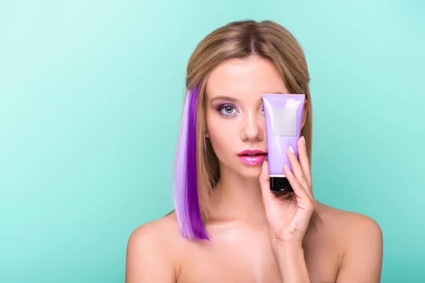 Hermosa mujer joven con pelo colorido sosteniendo el cabello tubo tónico y mirando a la cámara aislada en azul - foto de stock