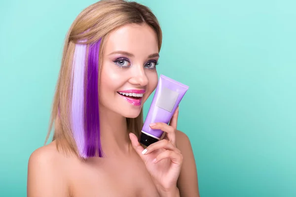 Happy young woman with colorful hair holding hair tonic tube and looking at camera isolated on blue — Stock Photo