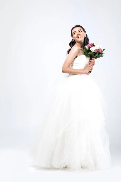 Hermosa novia joven en vestido de novia con ramo y mirando hacia otro lado en blanco - foto de stock