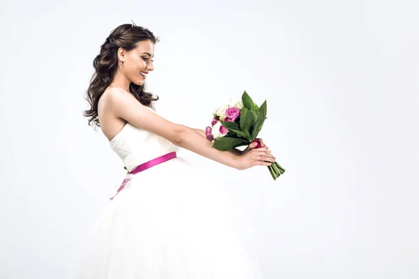 Vista lateral da jovem noiva sorridente em vestido de noiva segurando buquê de noiva isolado no branco — Fotografia de Stock