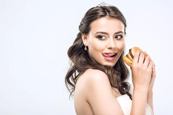 Hungry young bride in wedding dress with burger in hands showing tongue and looking away isolated on white — Stock Photo