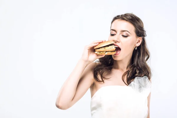 Hungry young bride in wedding dress eating big burger isolated on white — Stock Photo