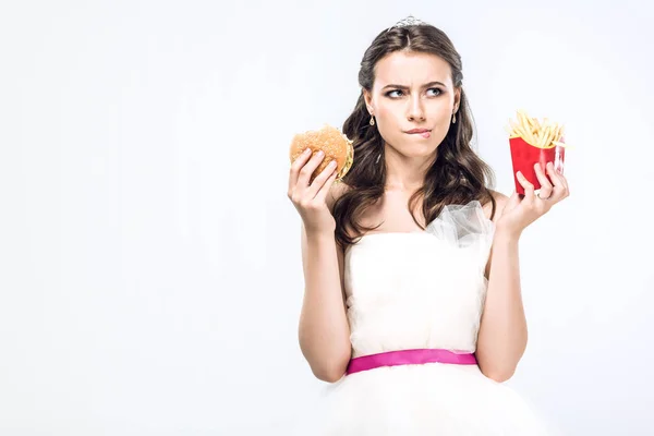 Reflexivo joven novia en vestido de novia con hamburguesa y papas fritas mirando hacia arriba aislado en blanco - foto de stock