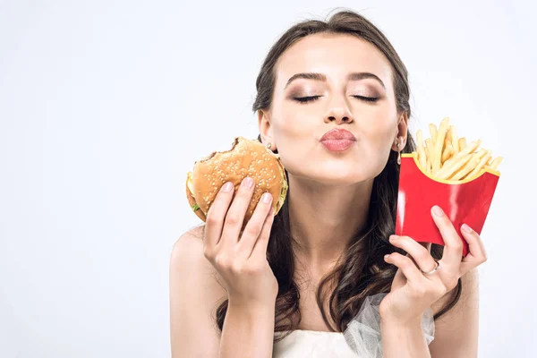 Joven novia en vestido de novia con hamburguesa y papas fritas enviando beso a cámara aislada en blanco - foto de stock