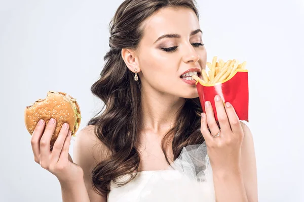 Young bride in wedding dress eating burger and french fries isolated on white — Stock Photo
