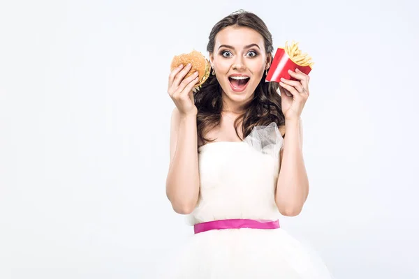 Shocked young bride in wedding dress with burger and french fries looking at camera isolated on white — Stock Photo