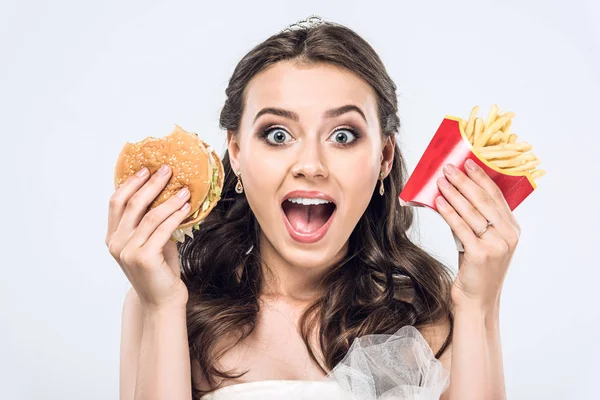 Portrait en gros plan de jeune mariée choquée en robe de mariée avec hamburger et frites en regardant la caméra isolée sur blanc — Photo de stock