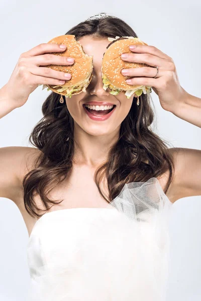 Laughing young bride in wedding dress covering eyes with burgers isolated on white — Stock Photo