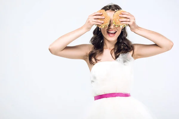 Smiling young bride in wedding dress covering eyes with burgers isolated on white — Stock Photo