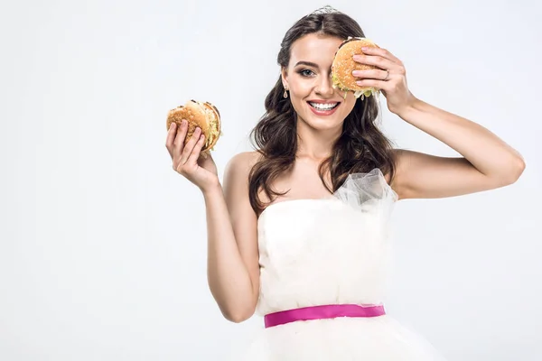 Sonriente joven novia en vestido de novia sosteniendo hamburguesas en manos aisladas en blanco - foto de stock
