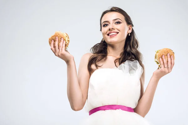 Vue du bas de la jeune mariée en robe de mariée tenant des hamburgers dans les mains isolées sur blanc — Photo de stock