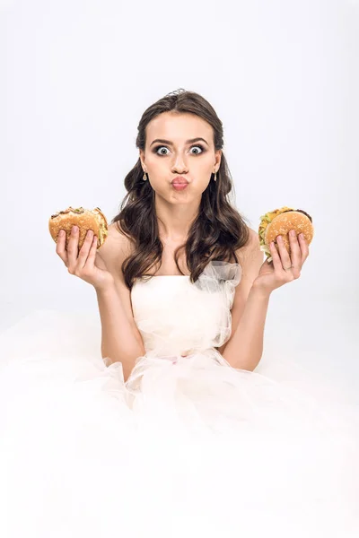 Attractive young bride in wedding dress holding burgers in hands while grimacing and looking at camera isolated on white — Stock Photo