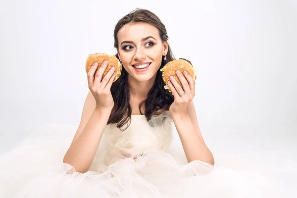Atractiva novia joven en vestido de novia sosteniendo hamburguesas en las manos y mirando hacia otro lado en blanco - foto de stock