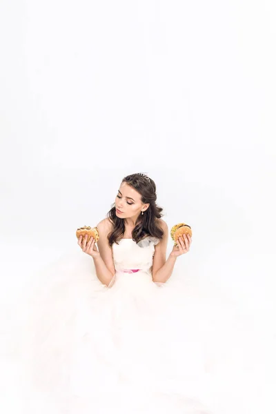 Attractive young bride in wedding dress sitting on floor with burgers in hands on white — Stock Photo