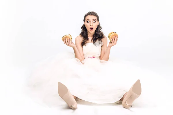 Shocked young bride in wedding dress sitting on floor with burgers in hands on white — Stock Photo
