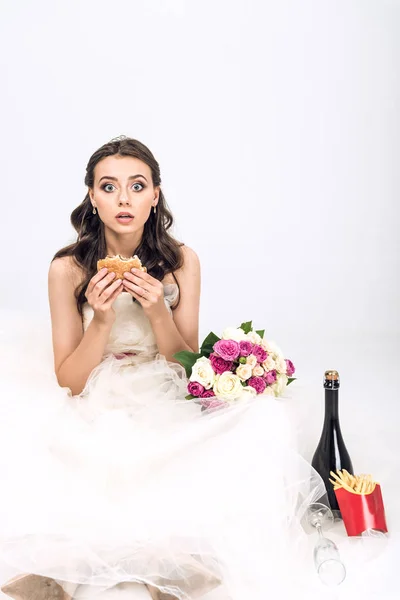 Shocked young bride in wedding dress sitting on floor with burger in hand on white — Stock Photo