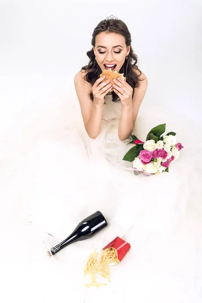 Visão de alto ângulo da noiva jovem no vestido de casamento comendo junk food enquanto sentado no chão no branco — Fotografia de Stock