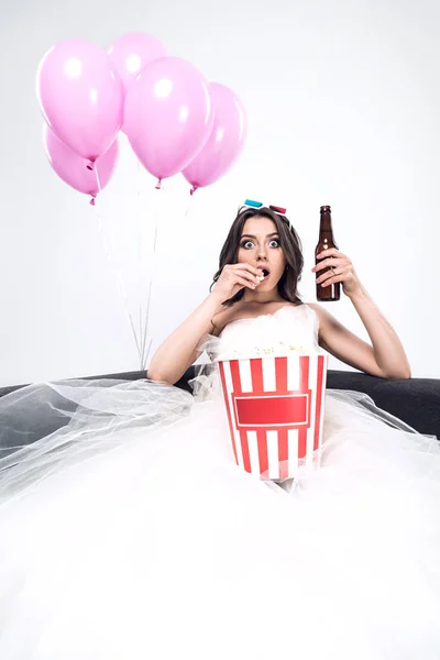 Shocked young bride in wedding dress with beer and bucket of popcorn watching movie isolated on white — Stock Photo