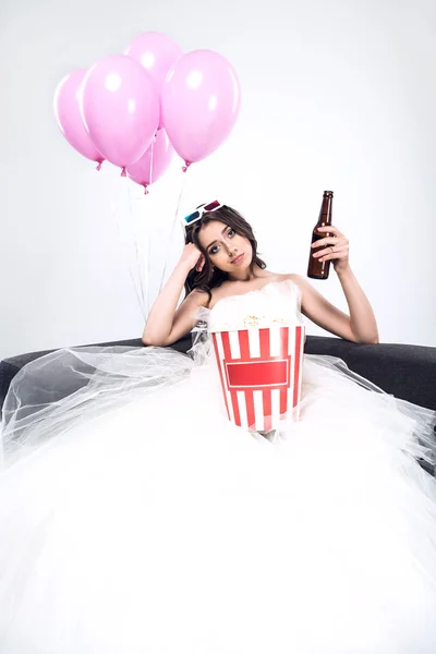 Confused young bride in wedding dress with beer and bucket of popcorn watching movie isolated on white — Stock Photo