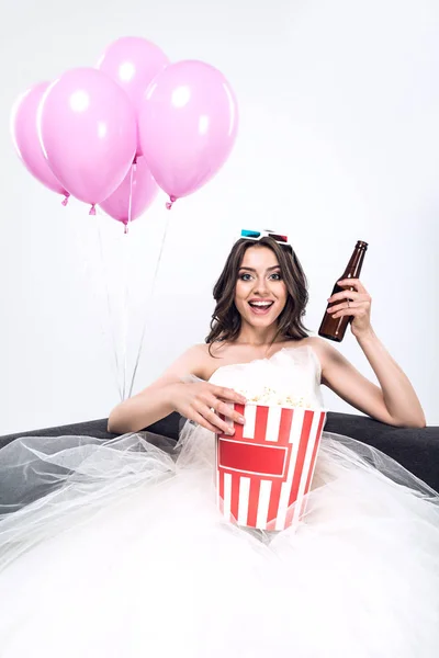 Happy young bride in wedding dress with beer and bucket of popcorn watching movie isolated on white — Stock Photo